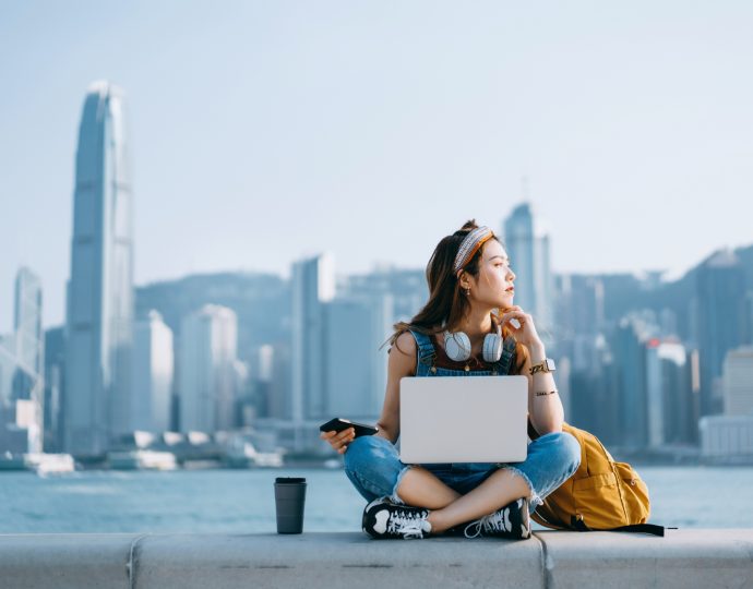 Beautiful young Asian woman sitting cross-legged by the promenade, against urban city skyline. She is wearing headphones around neck, using smartphone and working on laptop, with a coffee cup by her side. Looking away in thought. Lifestyle and technology